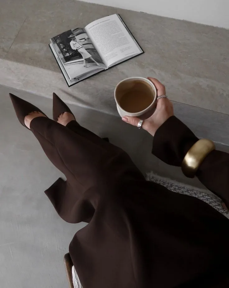 A woman in a cacao-brown suit holding a cup of coffee beside an open book