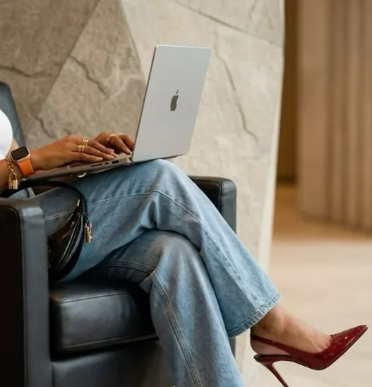 A woman working on a laptop in a leather armchair, in jeans and red heels